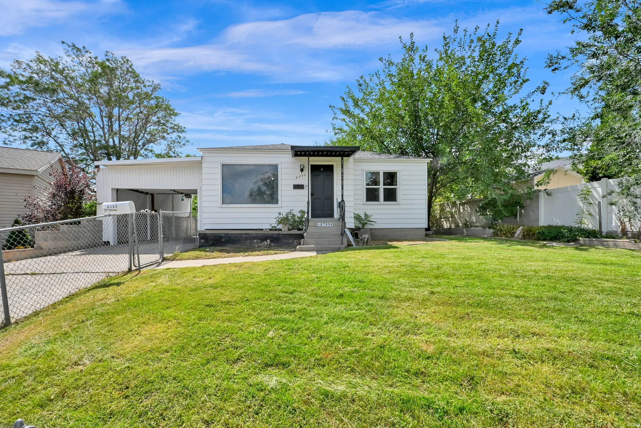 Single-story residence featuring white siding, a covered entryway, a front lawn, a chain-link fence, and a covered carport
