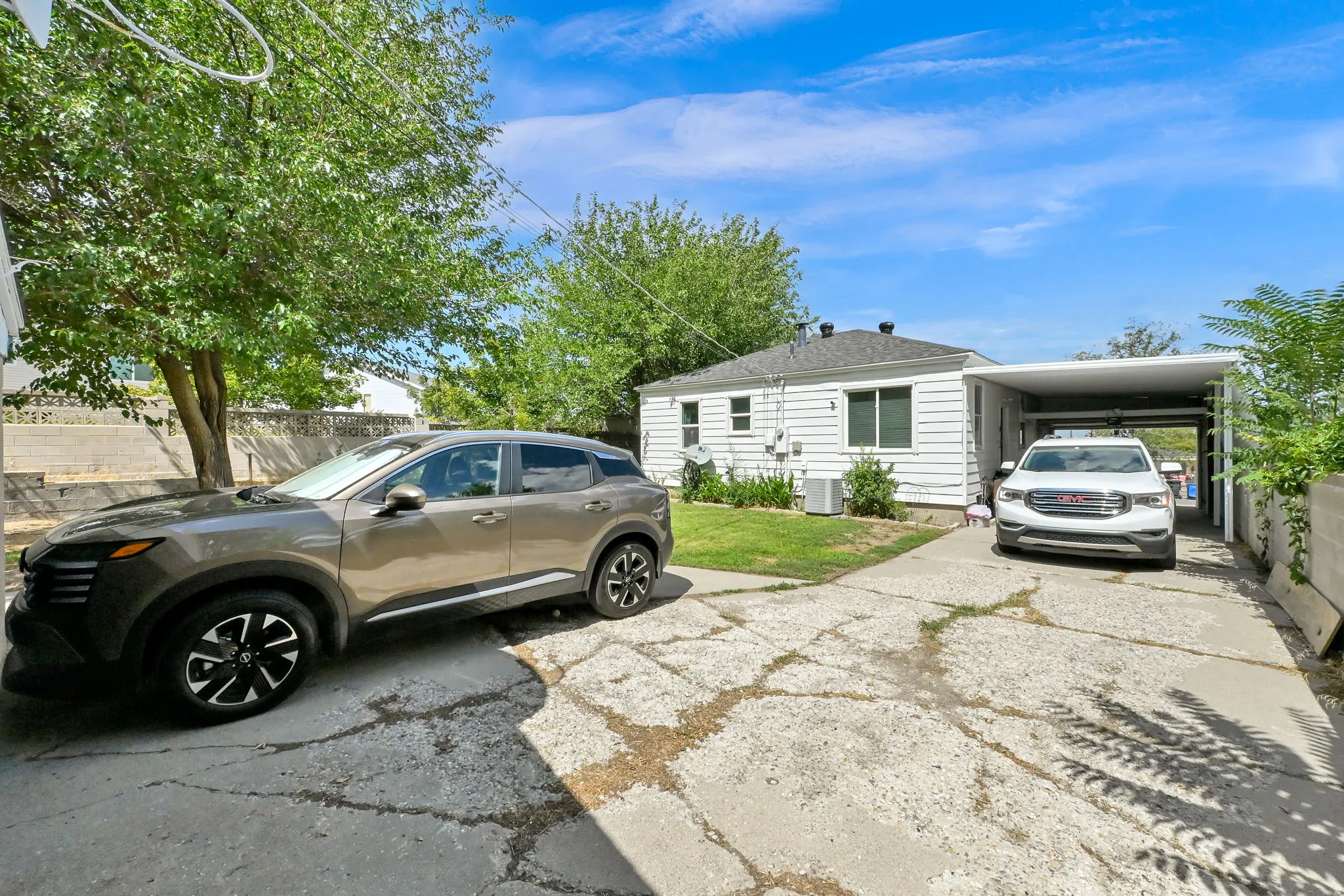 White siding exterior with a shaded carport, mature trees, and a concrete driveway