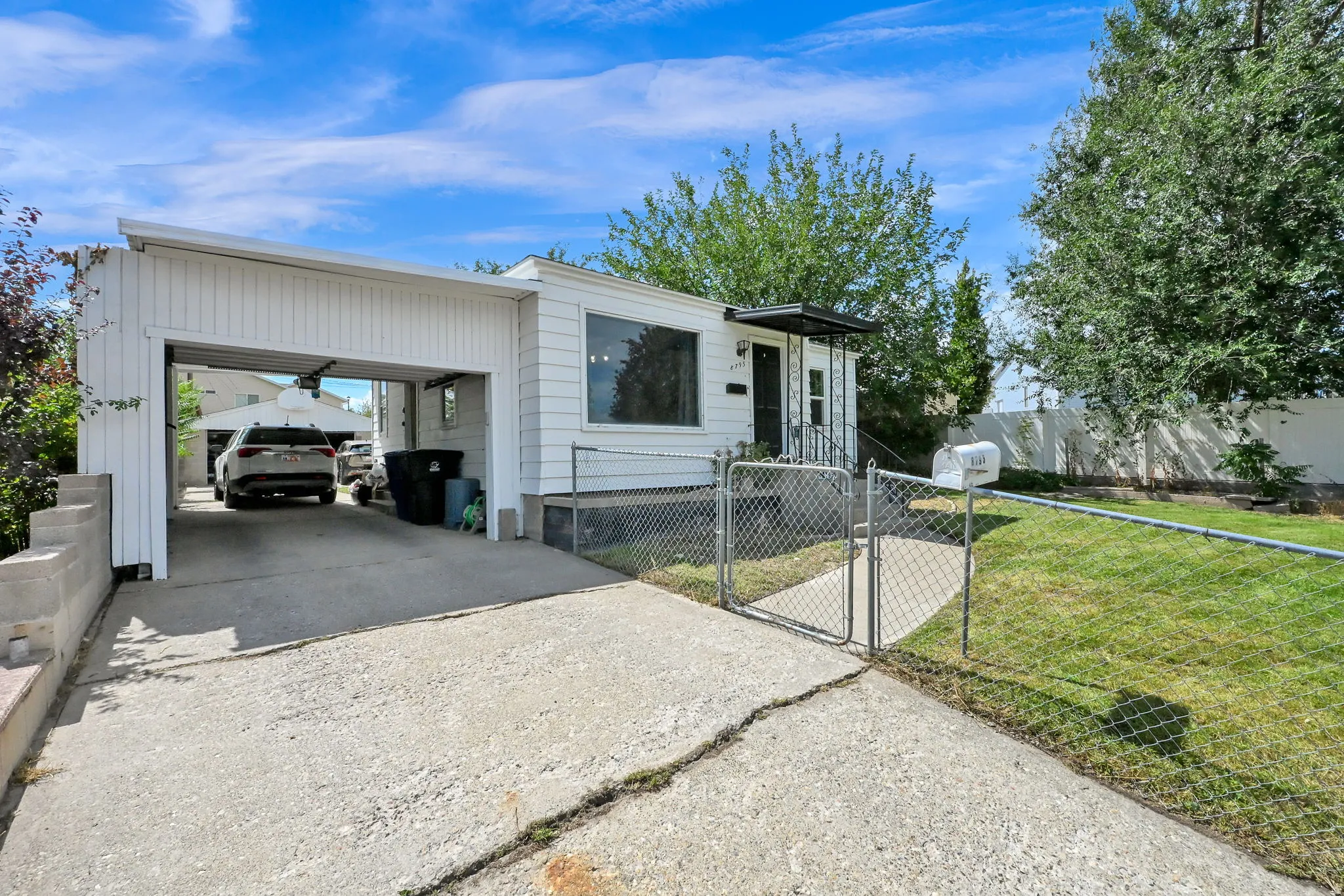 White siding exterior with a covered carport, concrete driveway, front entrance with an overhang, and a fenced front yard