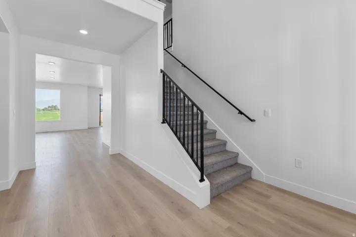 Light wood-finish flooring throughout, bright white walls, recessed ceiling lighting, a staircase with black metal railings, and a large window with exterior scenery