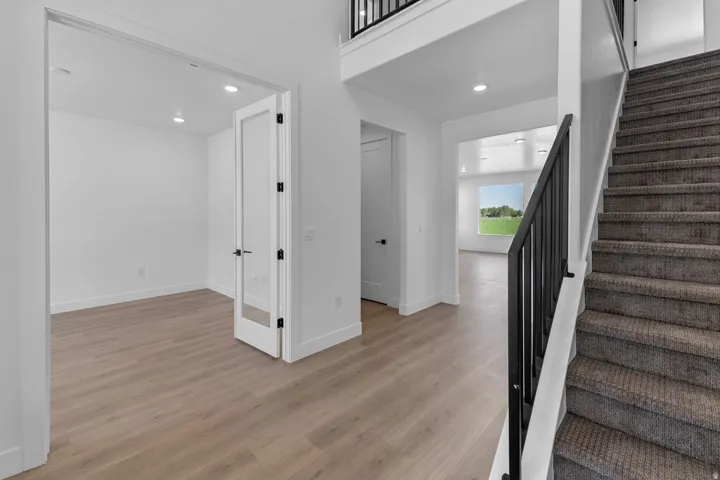 Open-concept interior featuring light wood-finish flooring, a carpeted staircase with a black metal railing, a solid white door, and a white interior door with a glass panel