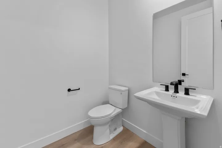 Pedestal sink with matte black fixtures, rectangular mirror, white toilet, and wood-finish flooring