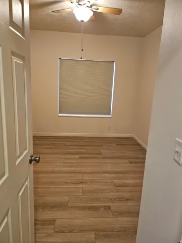 Wood-finish flooring, neutral wall tones, window with privacy blind, ceiling fan with integrated lighting, and white trim