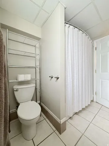 White toilet, tiled flooring, drop ceiling, and shower curtain