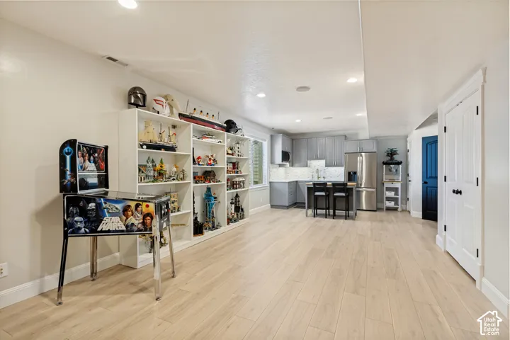 Recessed lighting illuminates the wood-finish flooring, built-in shelving, and a kitchen area featuring grey cabinetry, stainless steel appliances, and a central island with bar seating