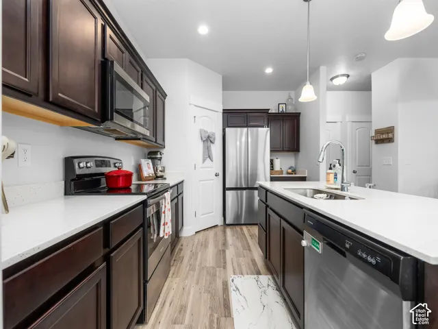 Kitchen featuring wood-finish flooring, dark wood cabinetry, stainless steel appliances, white countertops, and a kitchen island with an integrated sink