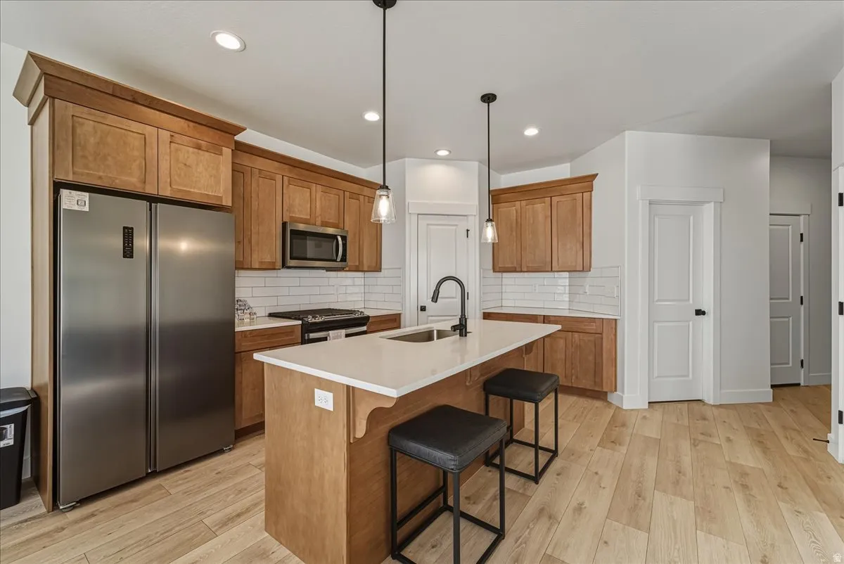 Kitchen featuring wood-finish flooring, stainless steel appliances, white subway tile backsplash, light wood cabinetry, and a center island with pendant lighting