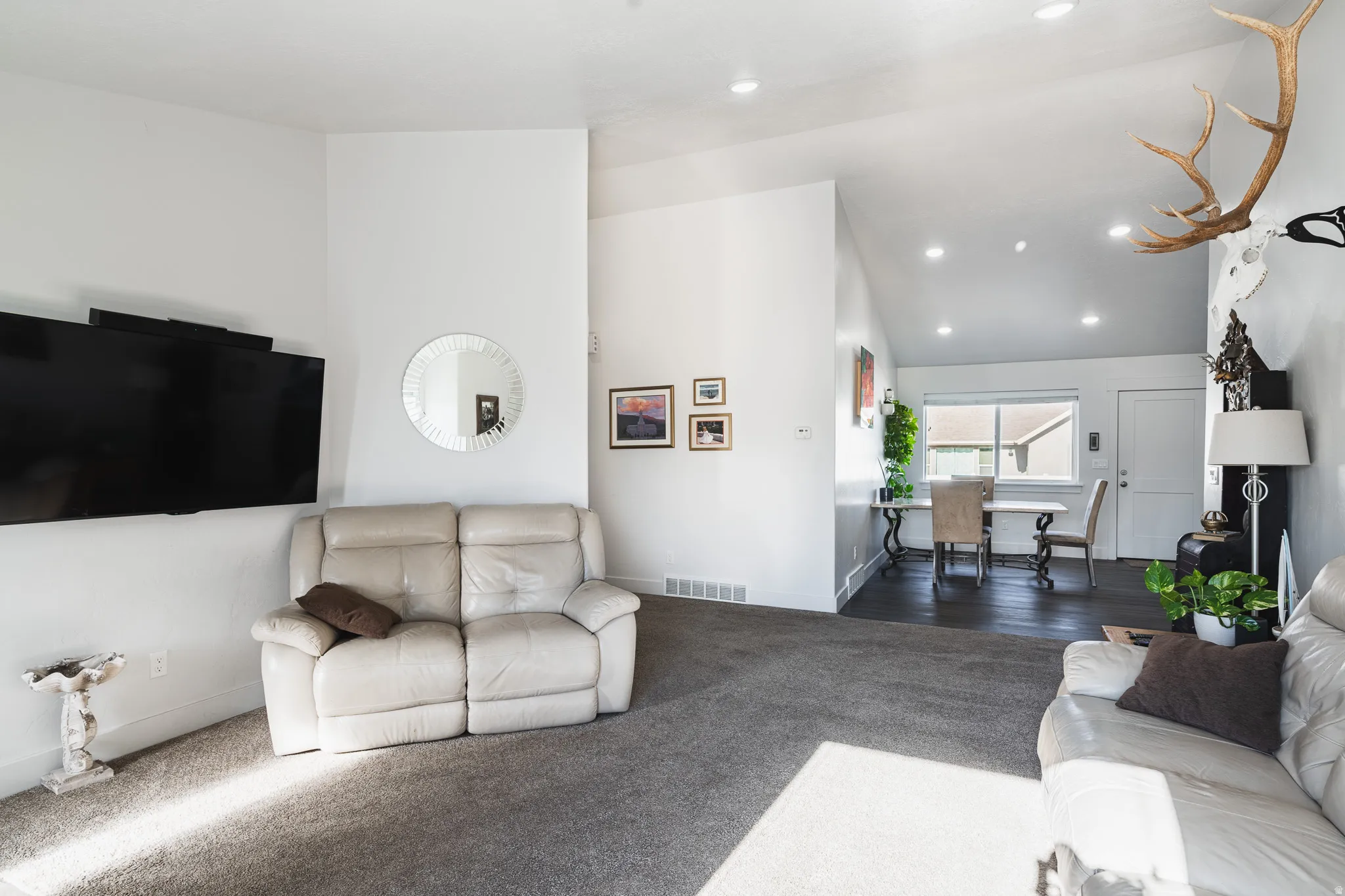 Recessed lighting illuminates the open-concept living space with vaulted ceilings, featuring a spacious carpeted area and dark wood-finish flooring extending into the dining area
