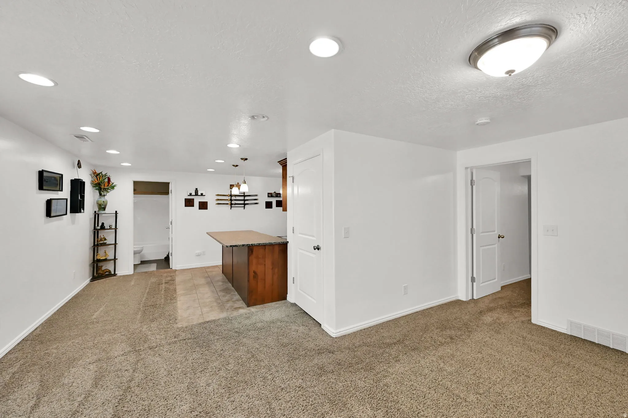 BASEMENT LIVING ROOM/KITCHEN. Recessed lighting throughout, wood-finish cabinetry with stone-finish countertop, neutral carpeting, and tile flooring