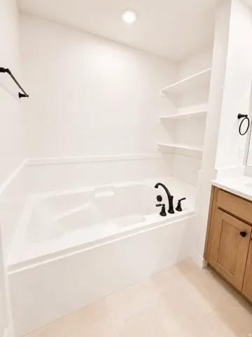 Built-in soaking tub with matte black fixtures, integrated shelving, and light-toned tile flooring