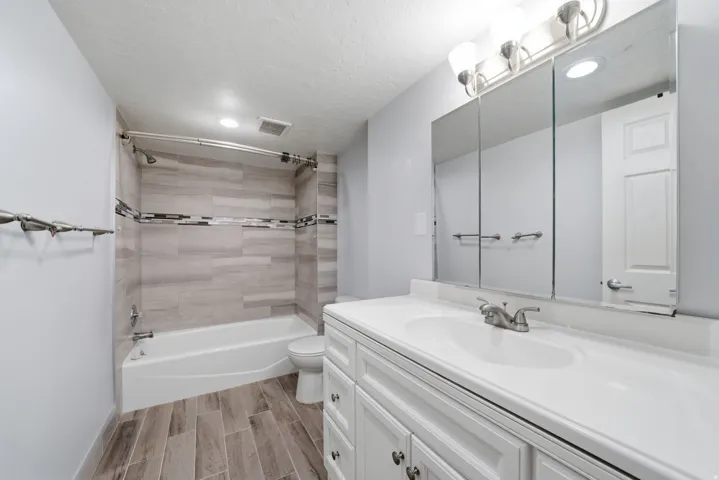 Bathroom featuring a white vanity with an integrated sink, a triple-door mirrored medicine cabinet, a three-light fixture, a shower/tub combination with patterned tile surround, and wood-finish flooring