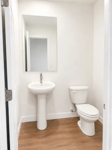 Pedestal sink with a polished chrome faucet, rectangular wall mirror, and wood-finish flooring