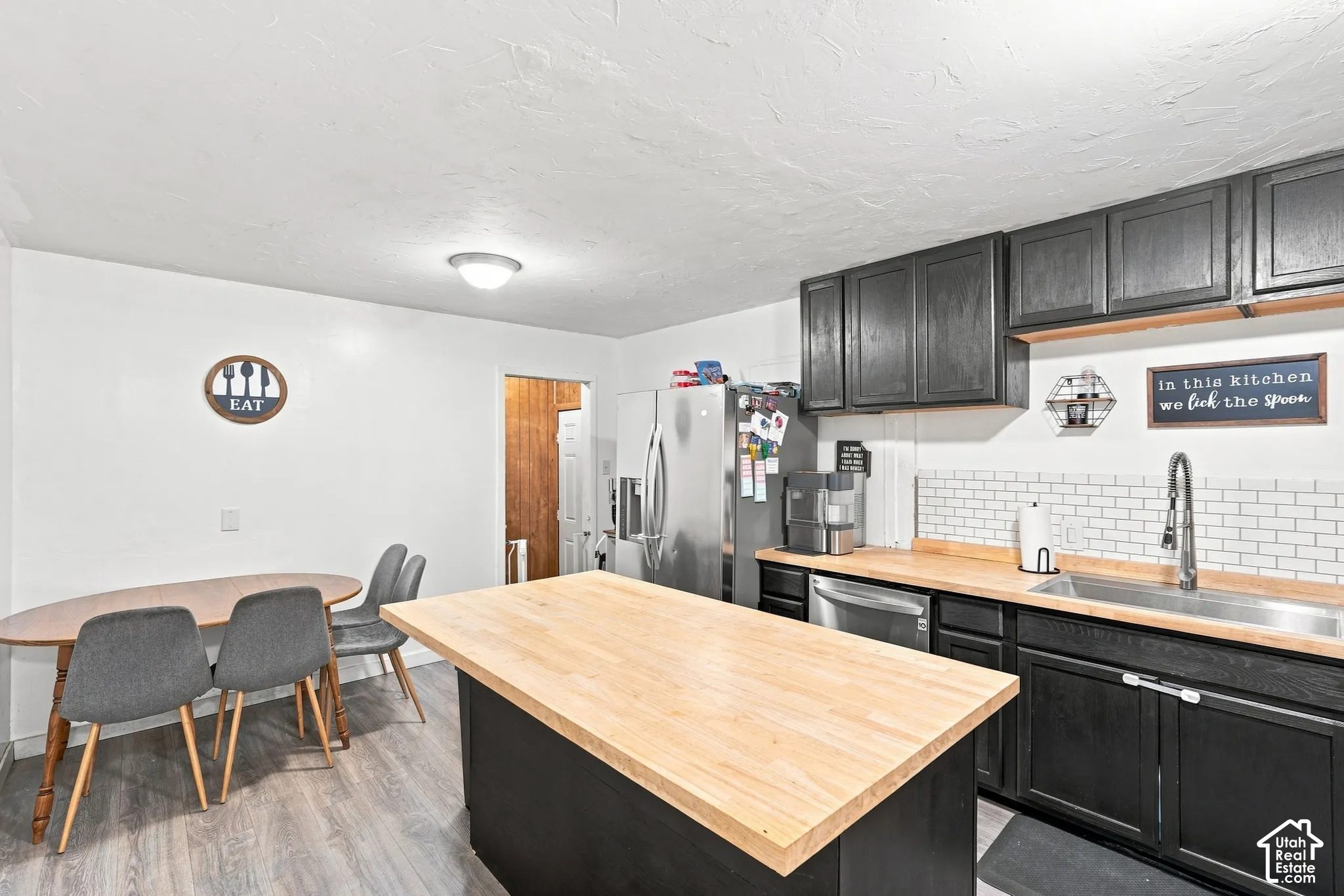 Kitchen featuring black cabinetry, butcher block countertops, subway tile backsplash, stainless steel refrigerator, and wood-finish flooring