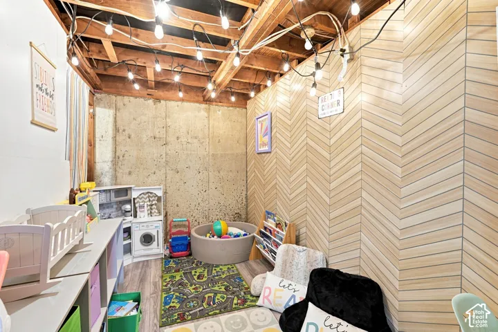 Exposed ceiling beams, string lighting, wood-finish chevron accent wall, concrete foundation wall, and wood-finish flooring