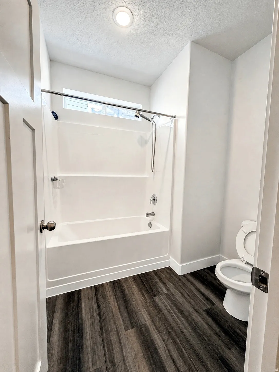 White tub and shower combination with a single window and wood-finish flooring