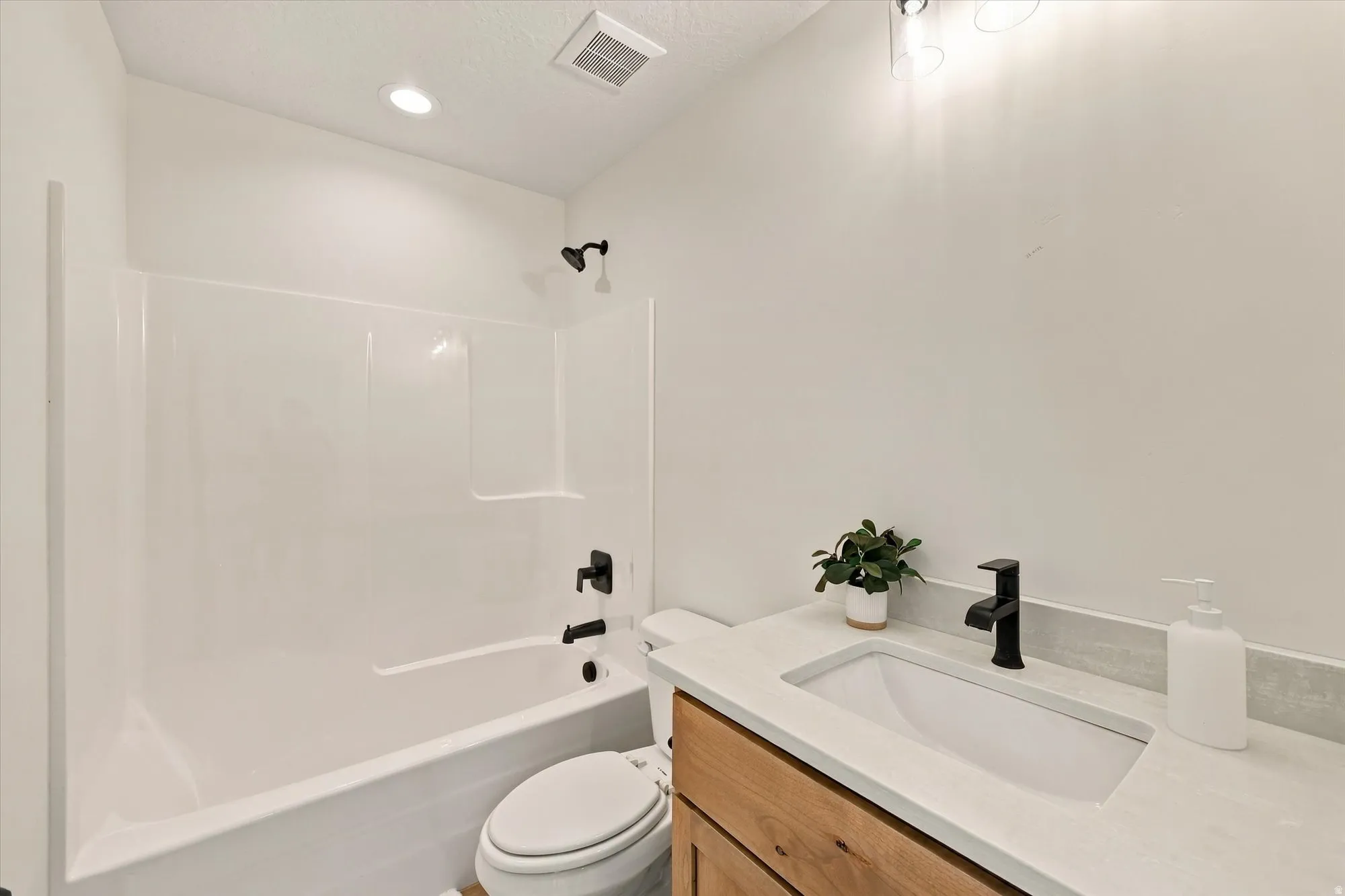 White bathtub with integrated shower, single-sink vanity with light-toned countertop, wood-finish cabinetry, matte black fixtures, and recessed ceiling lighting