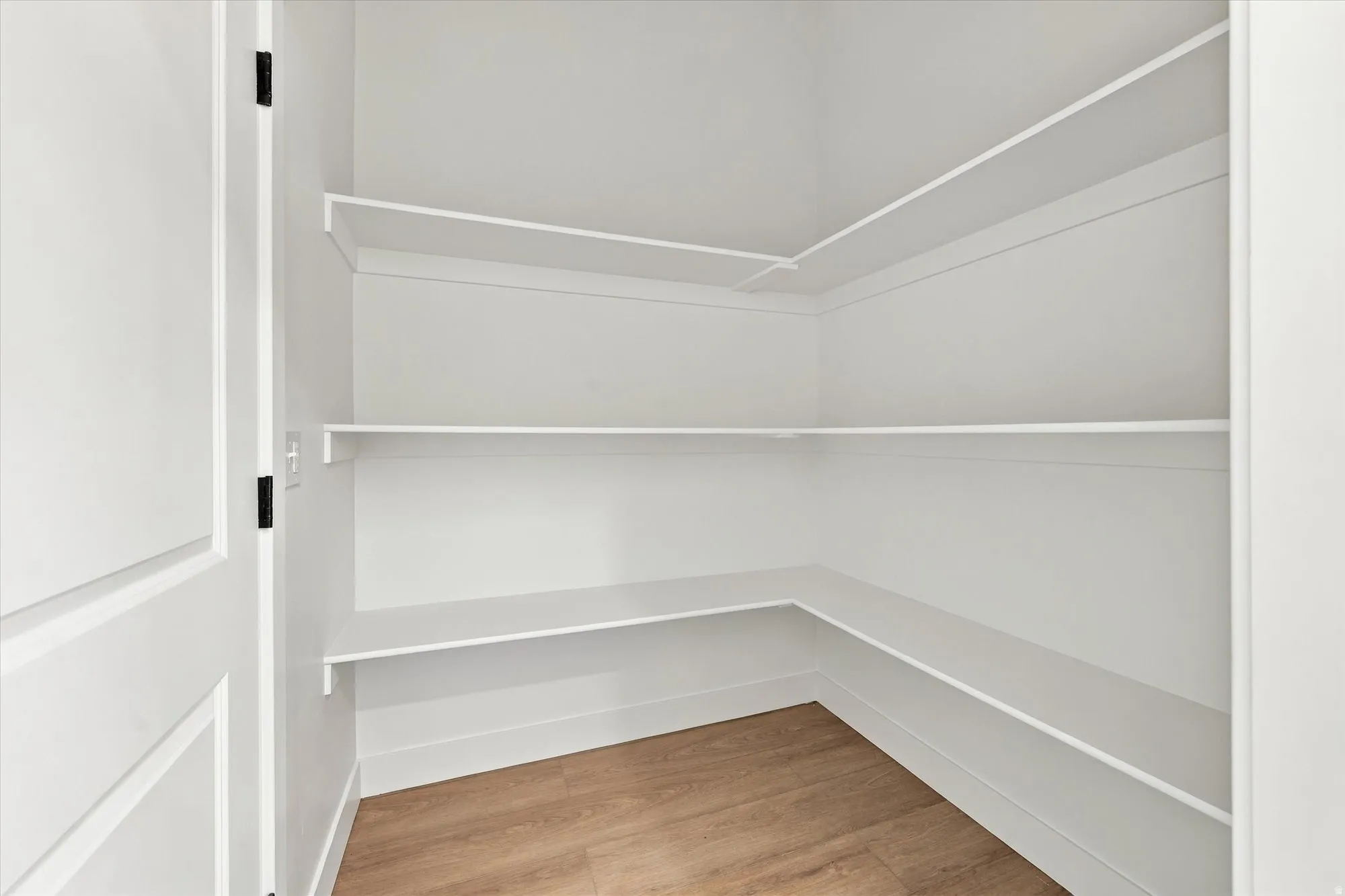 Walk-in pantry featuring light wood-finish flooring, white built-in shelving, and a white interior door with black hardware