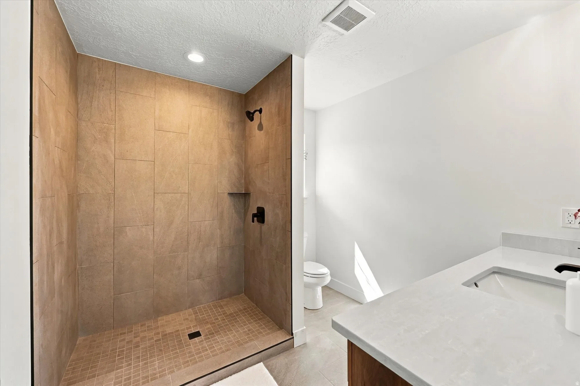 Walk-in shower with tiled walls and floor, recessed lighting, matte black shower fixtures, and a vanity with a white countertop and integrated sink