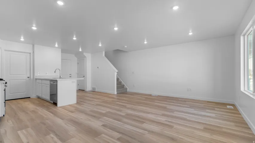 Open-concept living area featuring wood-finish flooring, recessed lighting, a kitchen island with a stainless steel sink, white cabinetry, and a carpeted staircase