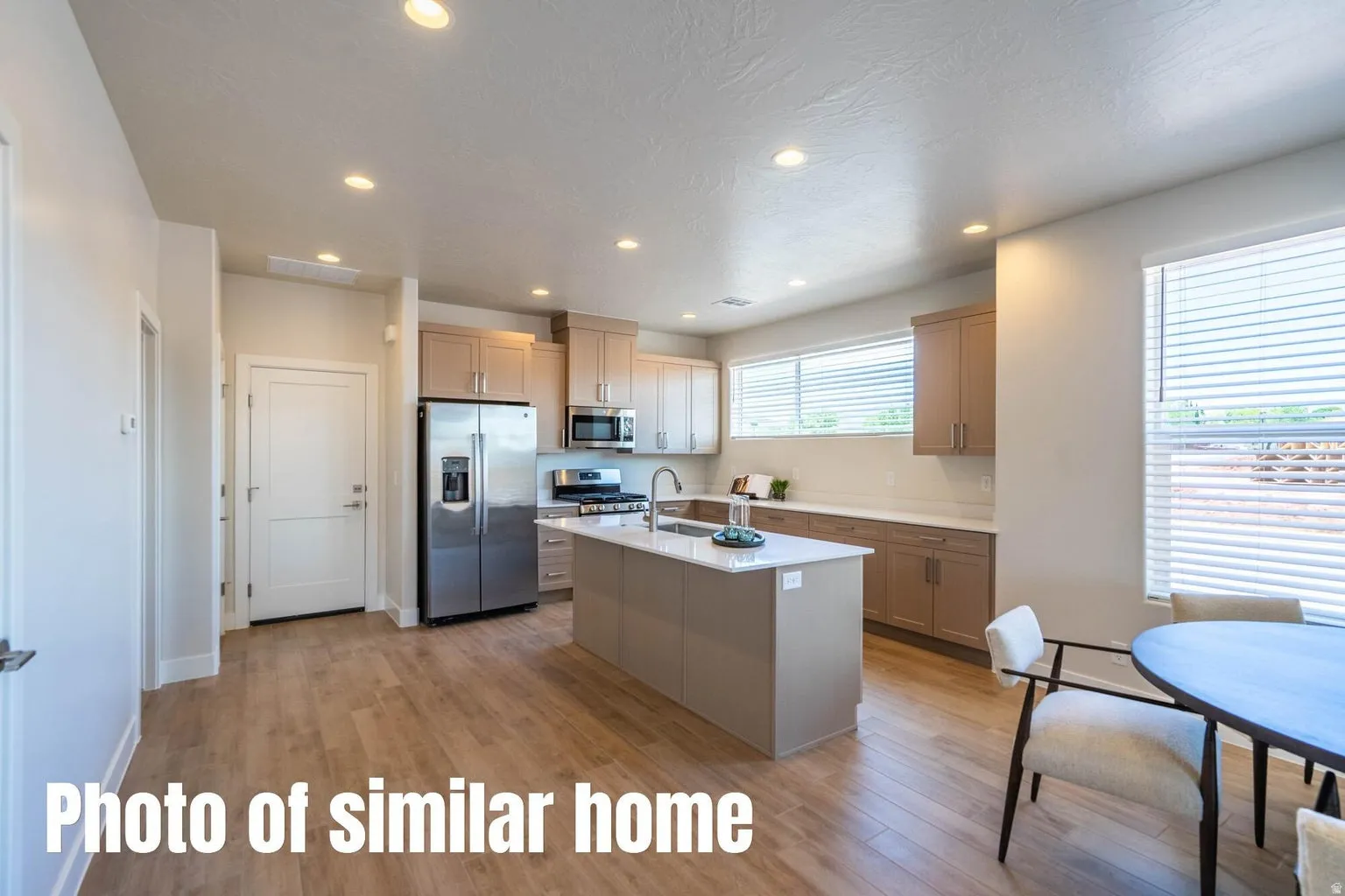 Modern kitchen featuring stainless steel appliances, white cabinetry with wood-finish upper accents, a large center island with seating space, quartz countertops, recessed ceiling lighting, wood-finish flooring throughout, and abundant natural light from