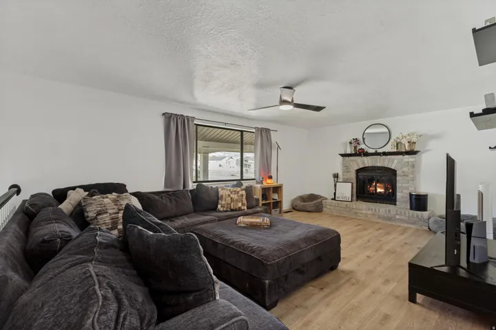 Wood-finish flooring, brick fireplace with a hearth, ceiling fan, and large window