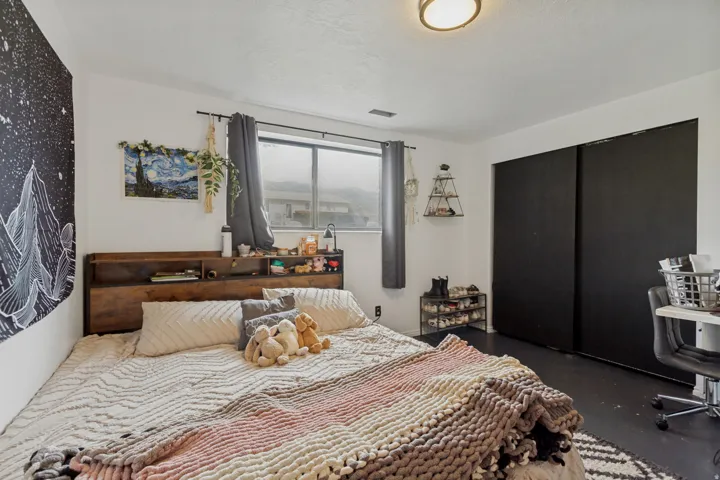 Recessed ceiling light fixture, window with roller shades, built-in wooden headboard with shelving, dark-toned flooring, and closet with sliding black doors