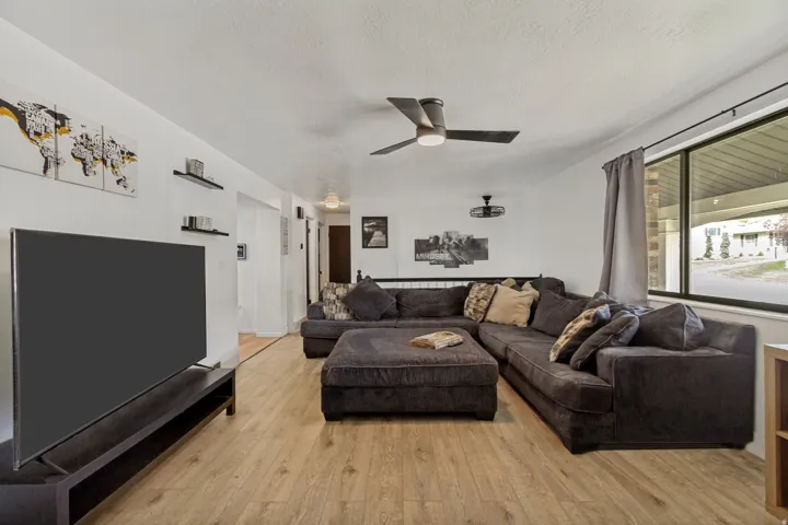White walls, wood-finish flooring, ceiling fan with integrated lighting, and a large window with privacy curtains