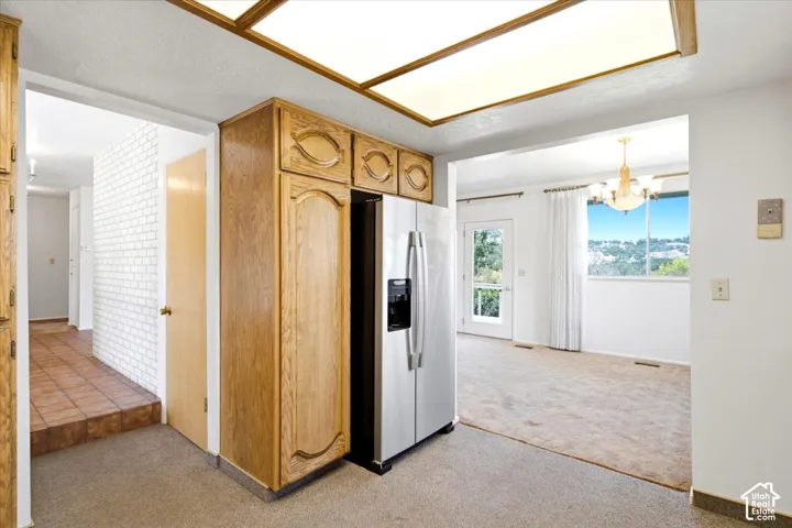 Kitchen features wood cabinetry and a side-by-side refrigerator, opening to a carpeted room with a chandelier and outdoor access via a glass-paneled door