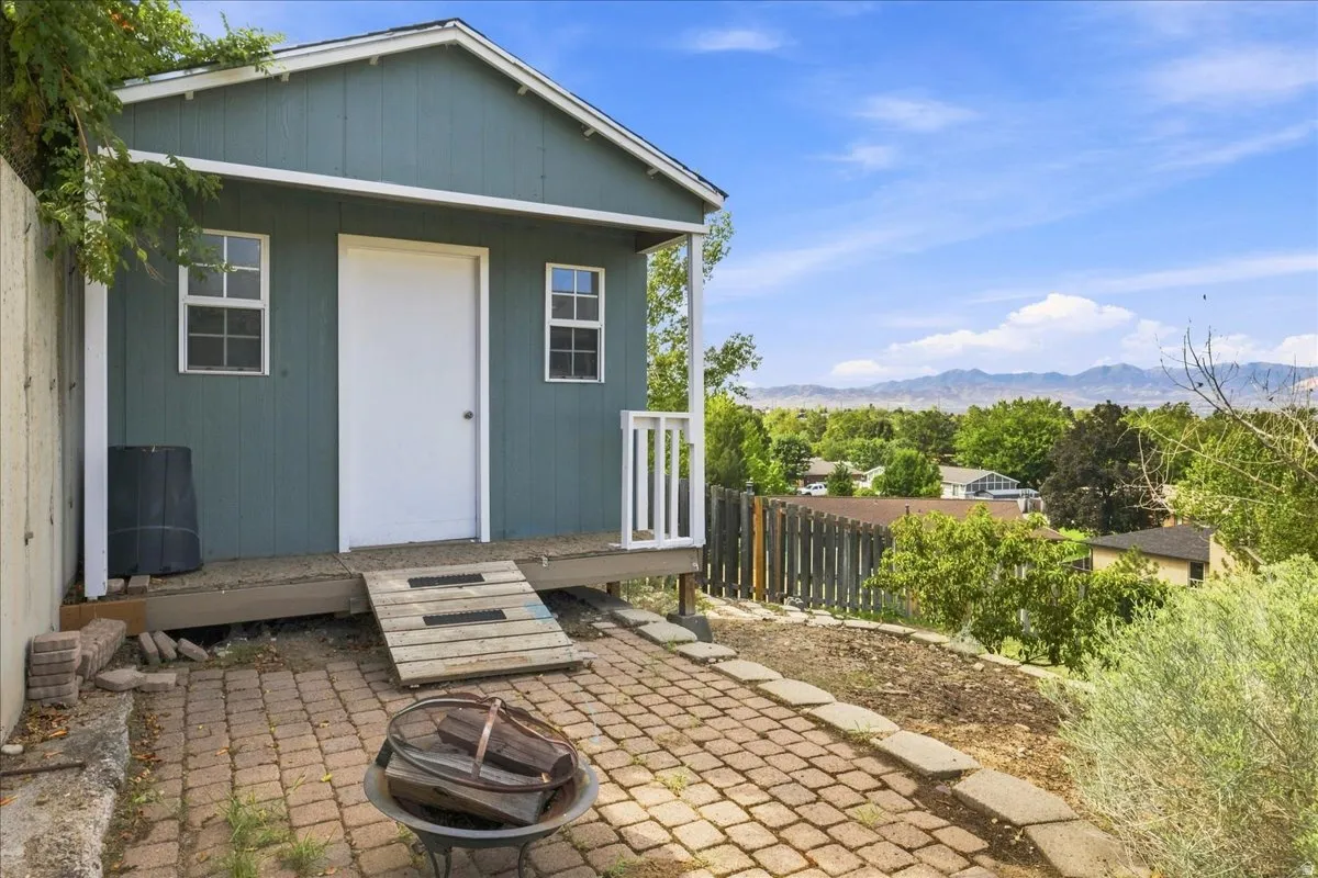 Freestanding outbuilding with a raised porch, two windows, and a solid door, featuring an adjacent brick paver patio and distant mountain scenery