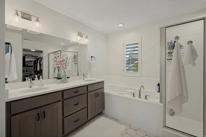 Dual vanity with white countertop and wood-finish cabinetry, built-in bathtub, glass-enclosed shower, plantation shutters, and hexagonal floor tiling