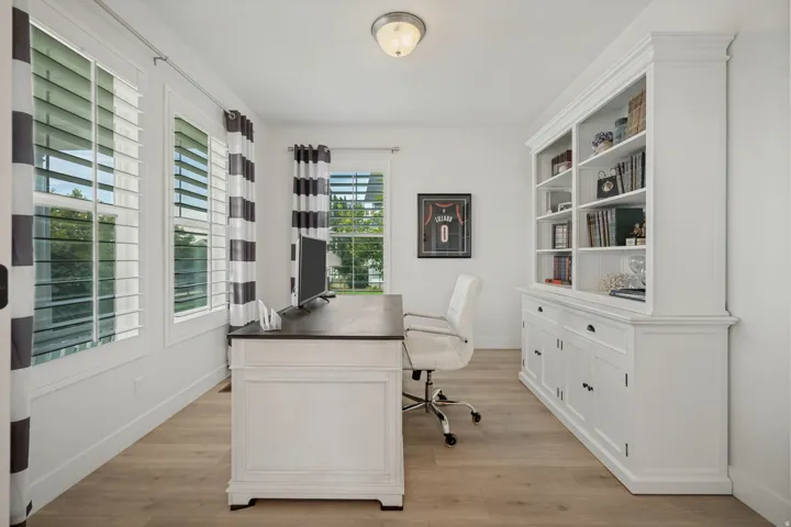 Bright interior with wood-finish flooring, multiple windows featuring plantation shutters, and a large built-in white cabinetry unit with shelving and lower storage