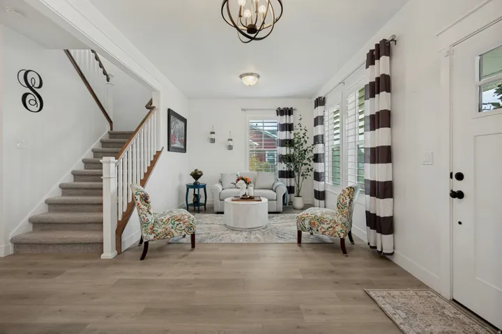 Open living area featuring wood-finish flooring, staircase with carpeted treads and white balusters, and white plantation shutters on multiple windows