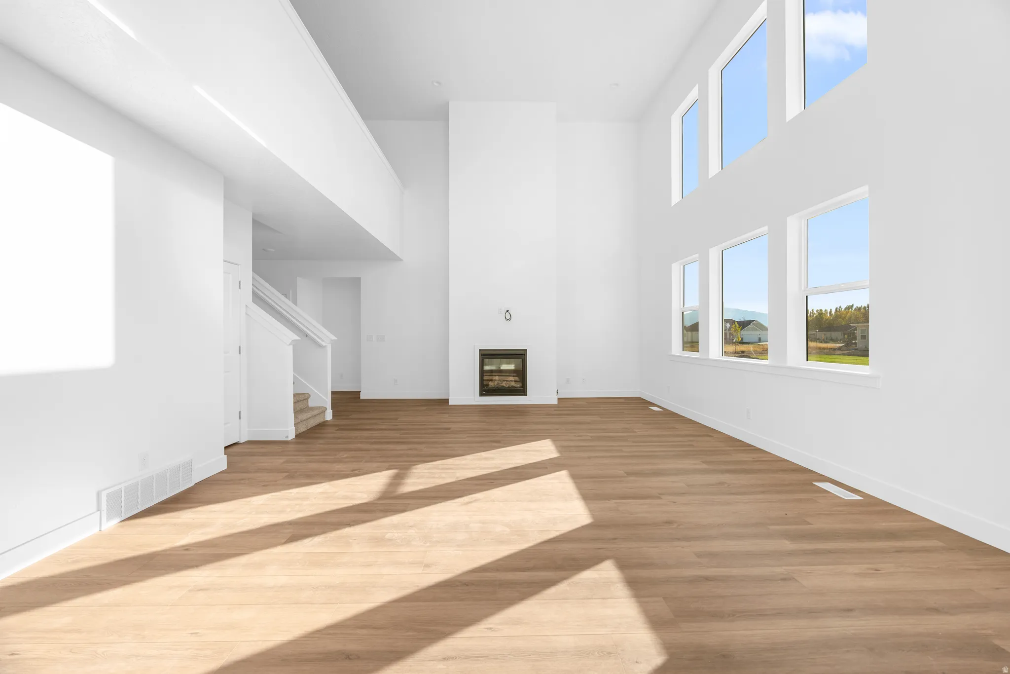 Soaring two-story ceilings with abundant natural light, a sleek linear fireplace, wood-finish flooring, and a white staircase with open railings