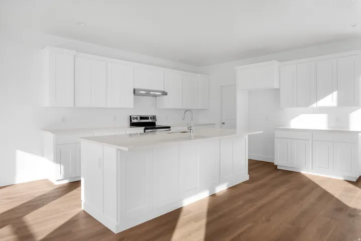 Bright kitchen featuring extensive white cabinetry, a large kitchen island with an integrated sink, light-toned countertops, stainless steel appliances, and wood-finish flooring