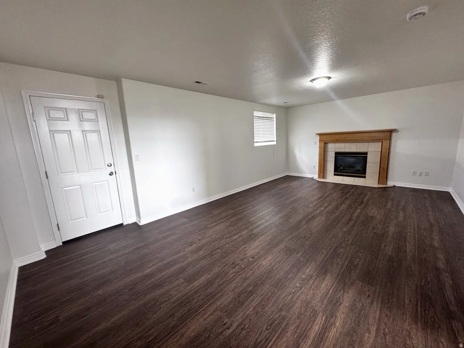 Spacious room featuring dark wood-finish flooring, a fireplace with a light wood mantel and tile surround, a window with horizontal blinds, and a white paneled door
