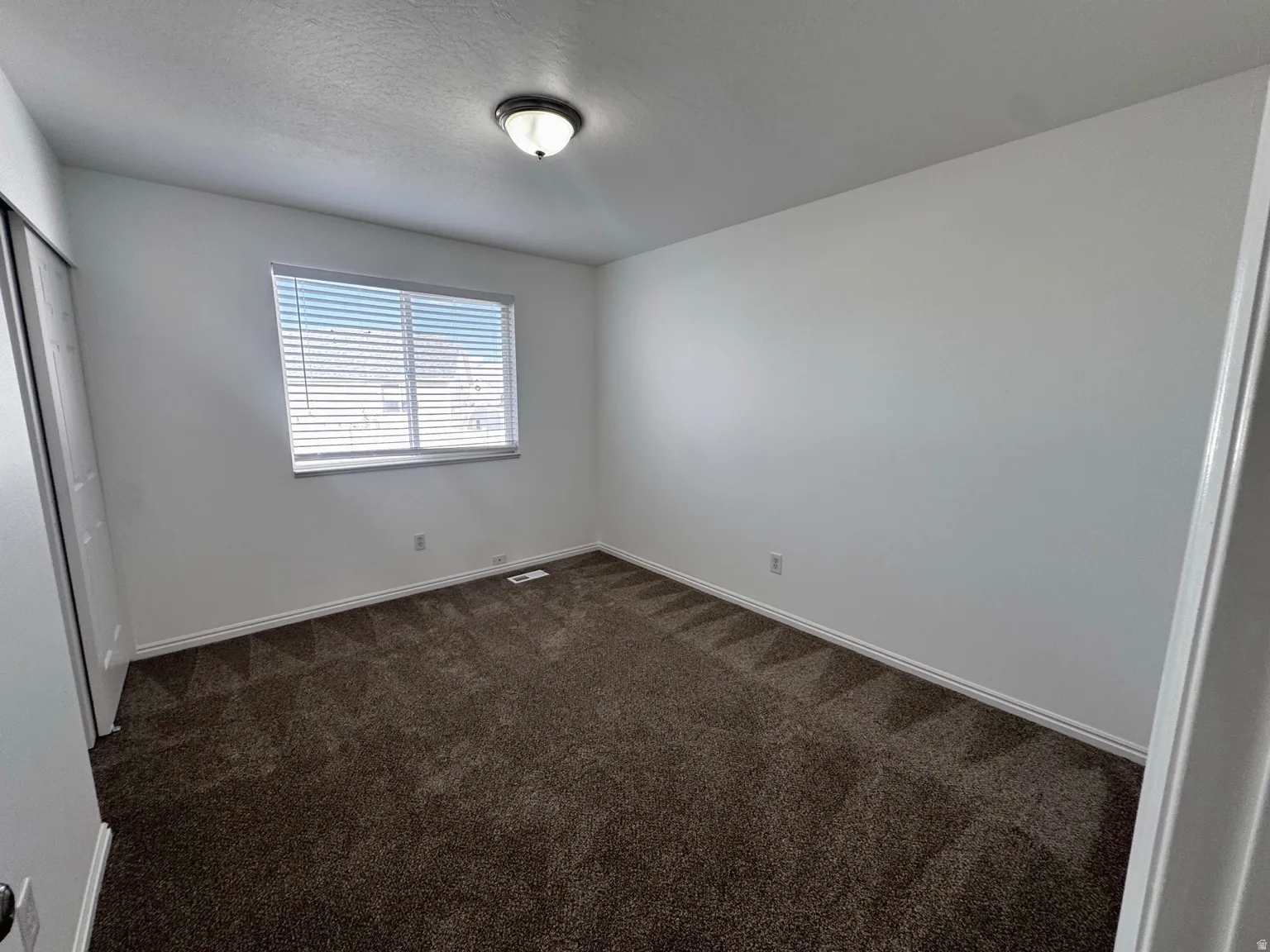Bright bedroom with expansive carpet flooring, large window with plantation shutters providing natural light, and clean white walls throughout