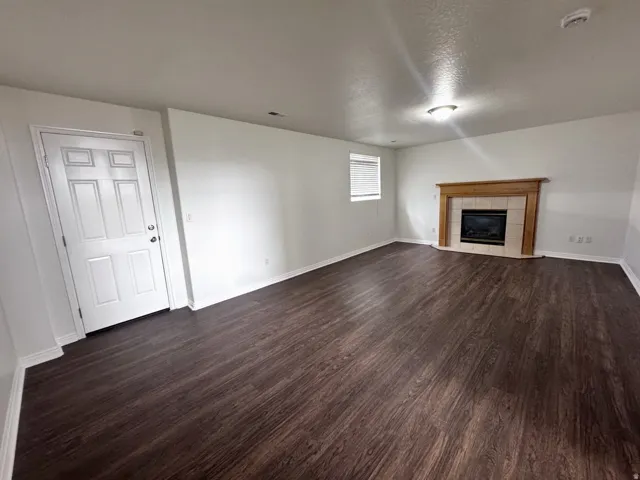 Spacious room featuring dark wood-finish flooring, a fireplace with a light wood mantel and tile surround, a window with horizontal blinds, and a white paneled door
