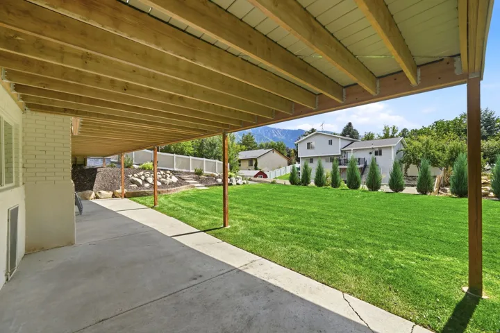 Covered concrete patio with exposed wood joist ceiling, a concrete path, a verdant lawn, and a tiered rock garden