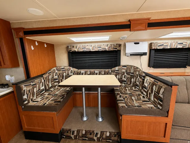 U-shaped dinette seating with inside travel trailer patterned upholstery, a light-toned table with dual pedestal bases, and two window blinds