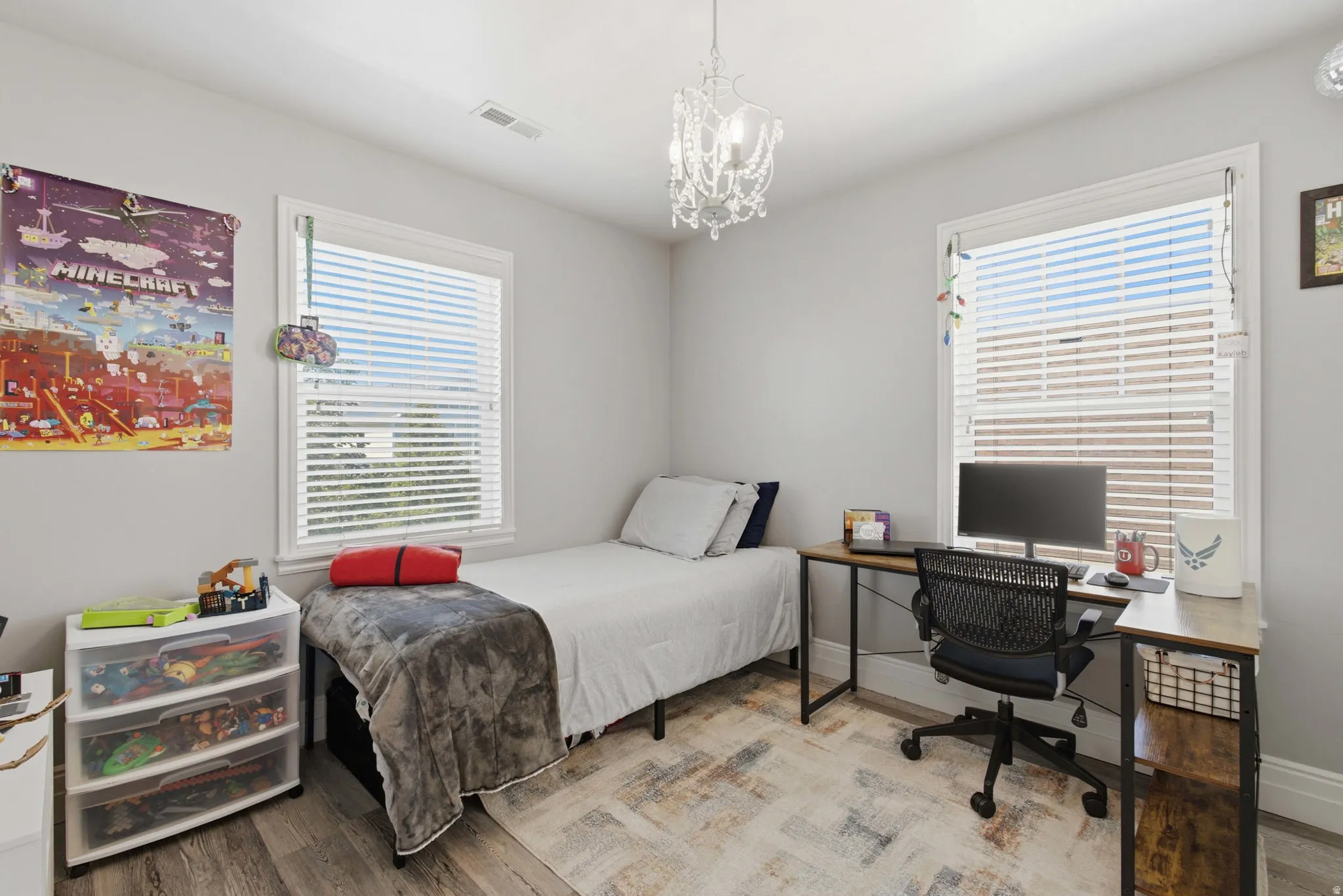Neutral wall color, wood-finish flooring, two large windows with blinds, and a decorative chandelier