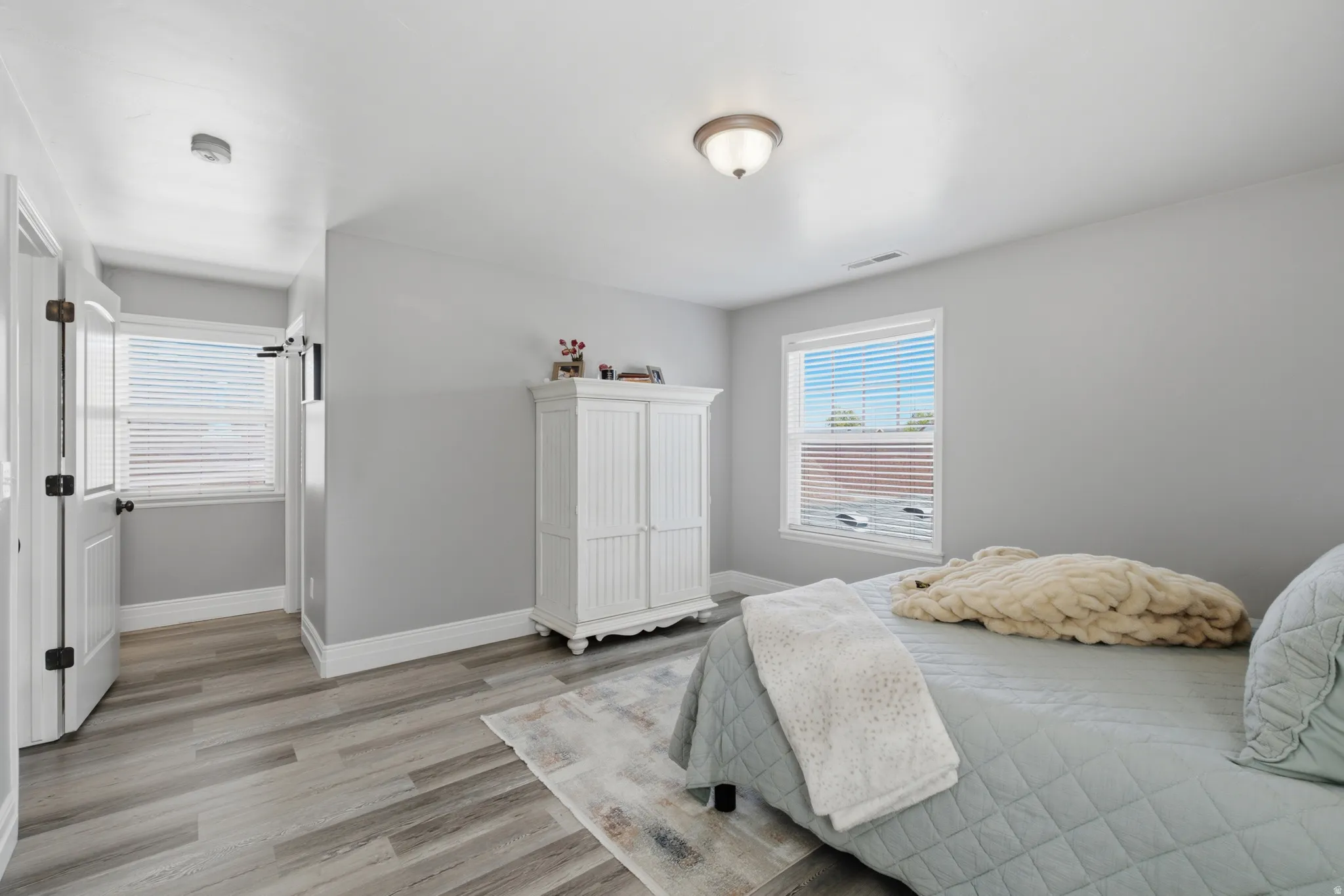 Owners Suite, Light gray walls with wood-finish flooring, featuring a window with horizontal blinds and a flush-mount ceiling light fixture