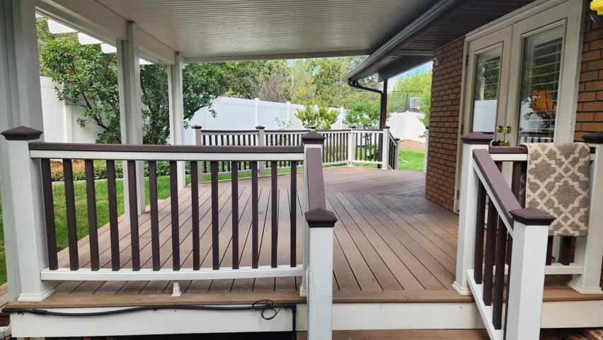 Expansive covered deck featuring composite decking, white railings with dark brown accents, and a brick exterior with double glass-paneled doors