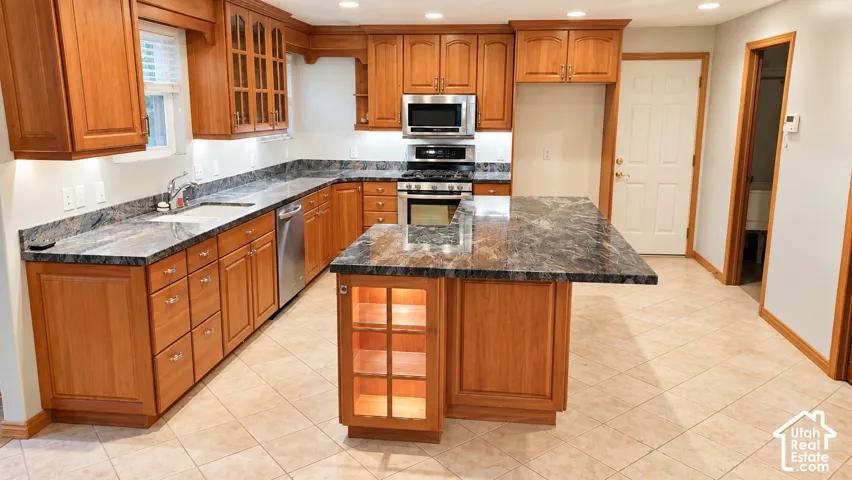 Kitchen featuring wood cabinetry, dark stone countertops, stainless steel appliances, a center island with illuminated display shelving, and tiled flooring