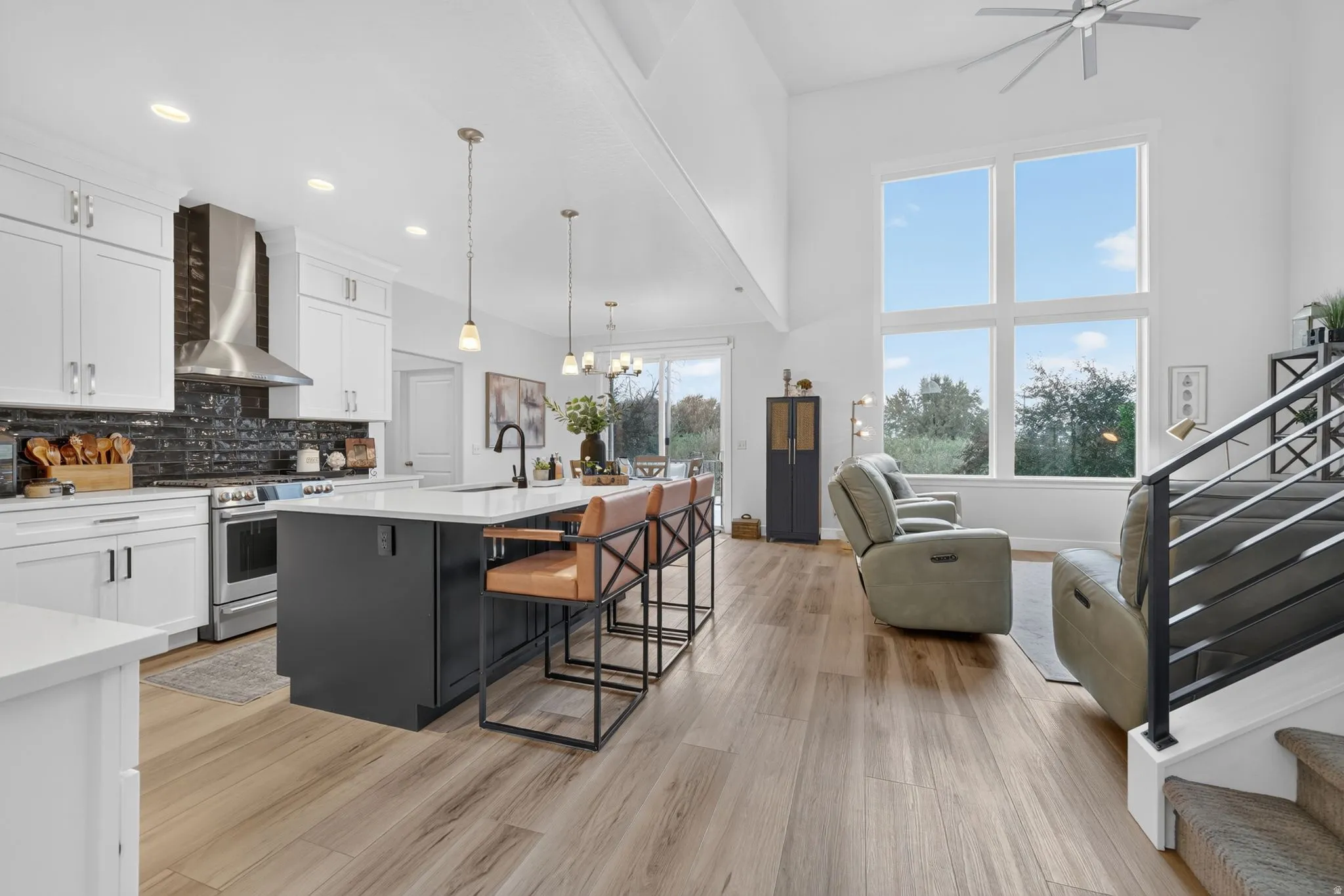 Open-concept interior featuring wood-finish flooring, white cabinetry, a kitchen island with seating, a stainless steel range hood, and a two-story window array