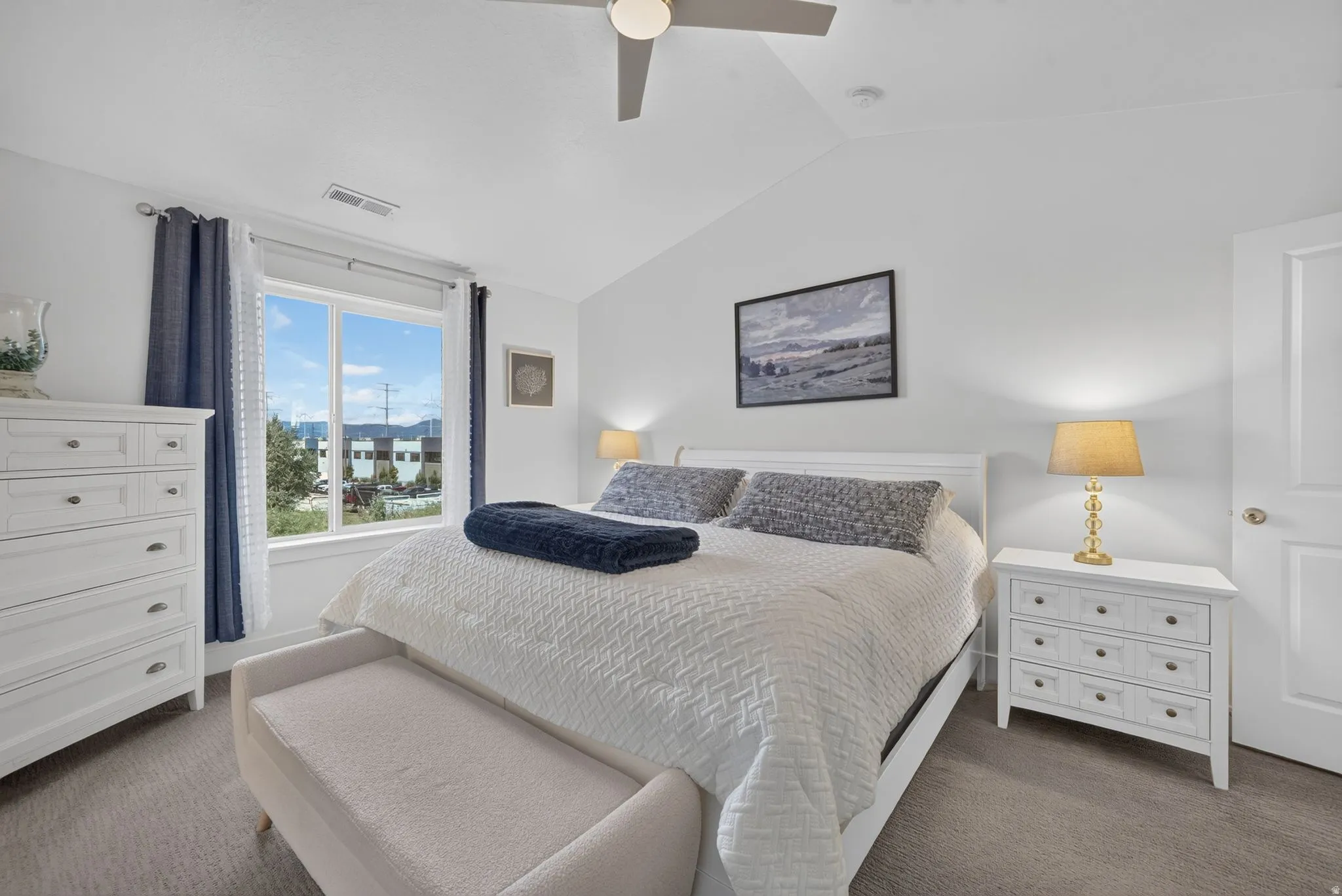 Bright, light-filled master bedroom featuring dual nightstands with brass table lamps, white cabinetry, ceiling fan, and expansive windows overlooking neighborhood streetscape