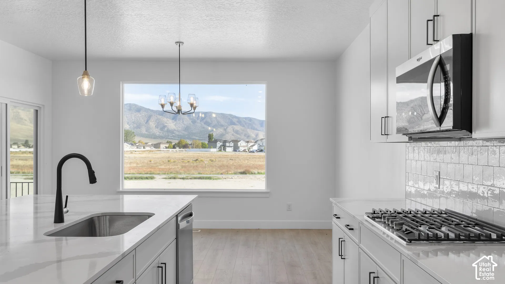 Open-concept kitchen featuring an integrated stainless steel sink, large window, pendant lighting, and light-toned cabinetry with dark hardware