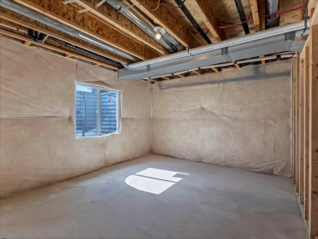 Unfinished basement with exposed ceiling joists, concrete floor, insulation-covered walls, visible ductwork, and a window well