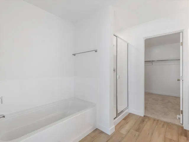 White bathtub with a chrome faucet, glass-enclosed shower, wood-finish flooring, and a walk-in closet with carpet