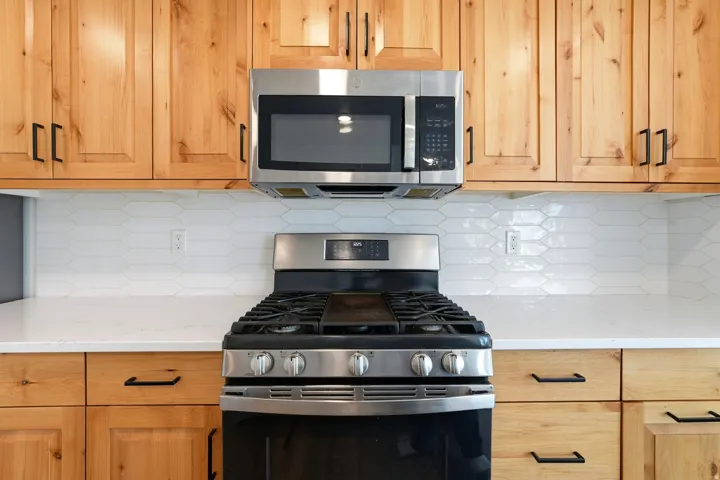 Wood cabinetry with black hardware, stainless steel range, built-in microwave, white countertops, and picket tile backsplash