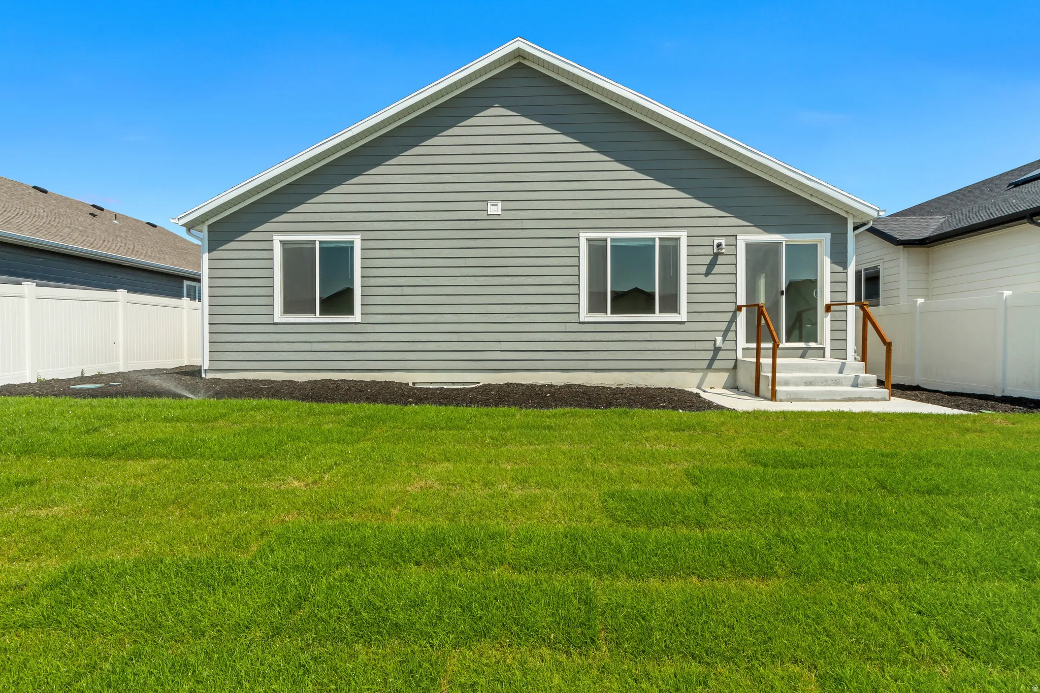 Spacious backyard with a vibrant green lawn, gray siding exterior, white vinyl fencing, and a concrete patio with steps